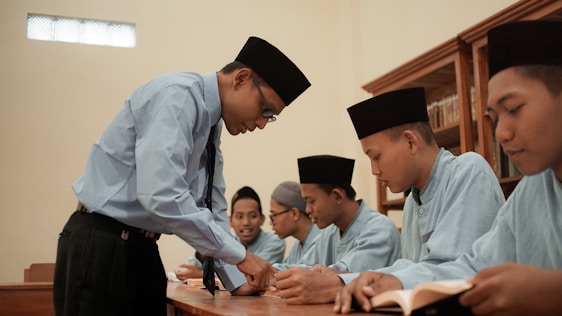 A teacher is engaging with a group of students sitting at a desk. The students are dressed in matching attire and wearing traditional headwear. They appear focused on their studies as they interact with the teacher. Books are open in front of them, and shelves filled with books are visible in the background.