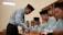 A teacher is engaging with a group of students sitting at a desk. The students are dressed in matching attire and wearing traditional headwear. They appear focused on their studies as they interact with the teacher. Books are open in front of them, and shelves filled with books are visible in the background.