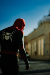 A person dressed in a dark superhero costume with red accents and a spider emblem on the back is standing on a street. The setting is urban, with an out-of-focus beige building in the background against a clear blue sky.