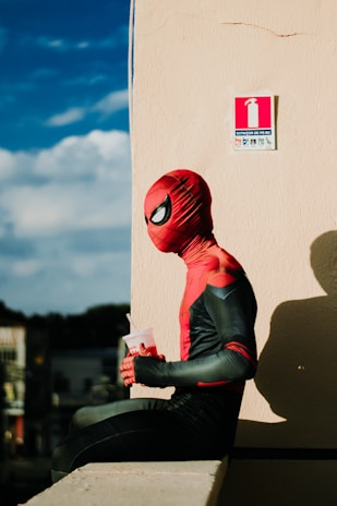 A person in a red and black superhero costume is sitting on a ledge. They are holding a drink cup with a straw. The background features a beige wall with a fire extinguisher sign and a blue sky with clouds.