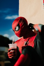 A person in a red and black superhero costume is sitting against a beige wall, holding a drink with a red straw. The costume is highly detailed with web-like patterns and a spider emblem on the chest. The lighting casts dramatic shadows, creating a contrast against the sunny background with a clear blue sky.