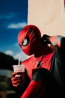 A person in a red and black superhero costume is sitting against a beige wall, holding a drink with a red straw. The costume is highly detailed with web-like patterns and a spider emblem on the chest. The lighting casts dramatic shadows, creating a contrast against the sunny background with a clear blue sky.