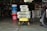 A garage is filled with various items including a stack of bags of Sakrete high-strength concrete mix placed on a yellow wheelbarrow. Additional bags are seen in the background. Nearby, a red bucket labeled 'Harbor Freight' stands next to several blue and white buckets. A bicycle tire is partially visible on the right side. The garage is dimly lit, with shelves and tools visible in the background.