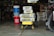 A garage is filled with various items including a stack of bags of Sakrete high-strength concrete mix placed on a yellow wheelbarrow. Additional bags are seen in the background. Nearby, a red bucket labeled 'Harbor Freight' stands next to several blue and white buckets. A bicycle tire is partially visible on the right side. The garage is dimly lit, with shelves and tools visible in the background.