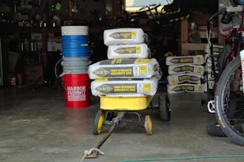 A garage is filled with various items including a stack of bags of Sakrete high-strength concrete mix placed on a yellow wheelbarrow. Additional bags are seen in the background. Nearby, a red bucket labeled 'Harbor Freight' stands next to several blue and white buckets. A bicycle tire is partially visible on the right side. The garage is dimly lit, with shelves and tools visible in the background.