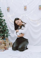 A woman kneels on a white fabric floor holding a black and white cat in her arms. She wears a white sweater and black pants, smiling gently. Around her are wrapped gifts and a decorated Christmas tree adorned with ornaments and lights. The background is draped with white fabric and small lights, contributing to a festive atmosphere.