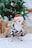 A small dog wearing a black and white cow-patterned costume and a red and green Santa hat stands in front of a decorated Christmas tree. Several wrapped gifts with ribbons and bows are placed on the floor around the dog. The scene is festive with holiday decorations including ornaments and snowflake designs on the tree.