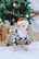 A small dog wearing a black and white cow-patterned costume and a red and green Santa hat stands in front of a decorated Christmas tree. Several wrapped gifts with ribbons and bows are placed on the floor around the dog. The scene is festive with holiday decorations including ornaments and snowflake designs on the tree.