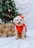 A small fluffy dog wearing a red outfit is sitting on a white cloth in front of a decorated Christmas tree. The tree is adorned with lights and ornaments, and there are several wrapped gifts with bows around the dog.