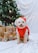 A small fluffy dog wearing a red outfit is sitting on a white cloth in front of a decorated Christmas tree. The tree is adorned with lights and ornaments, and there are several wrapped gifts with bows around the dog.