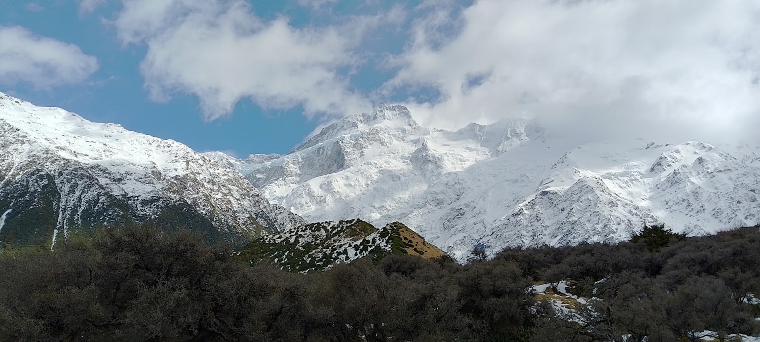 A large snowy mountain range half concealed by clouds. Some of the blue sky can be seen behind the mountains, and there are shrubs in the foreground.