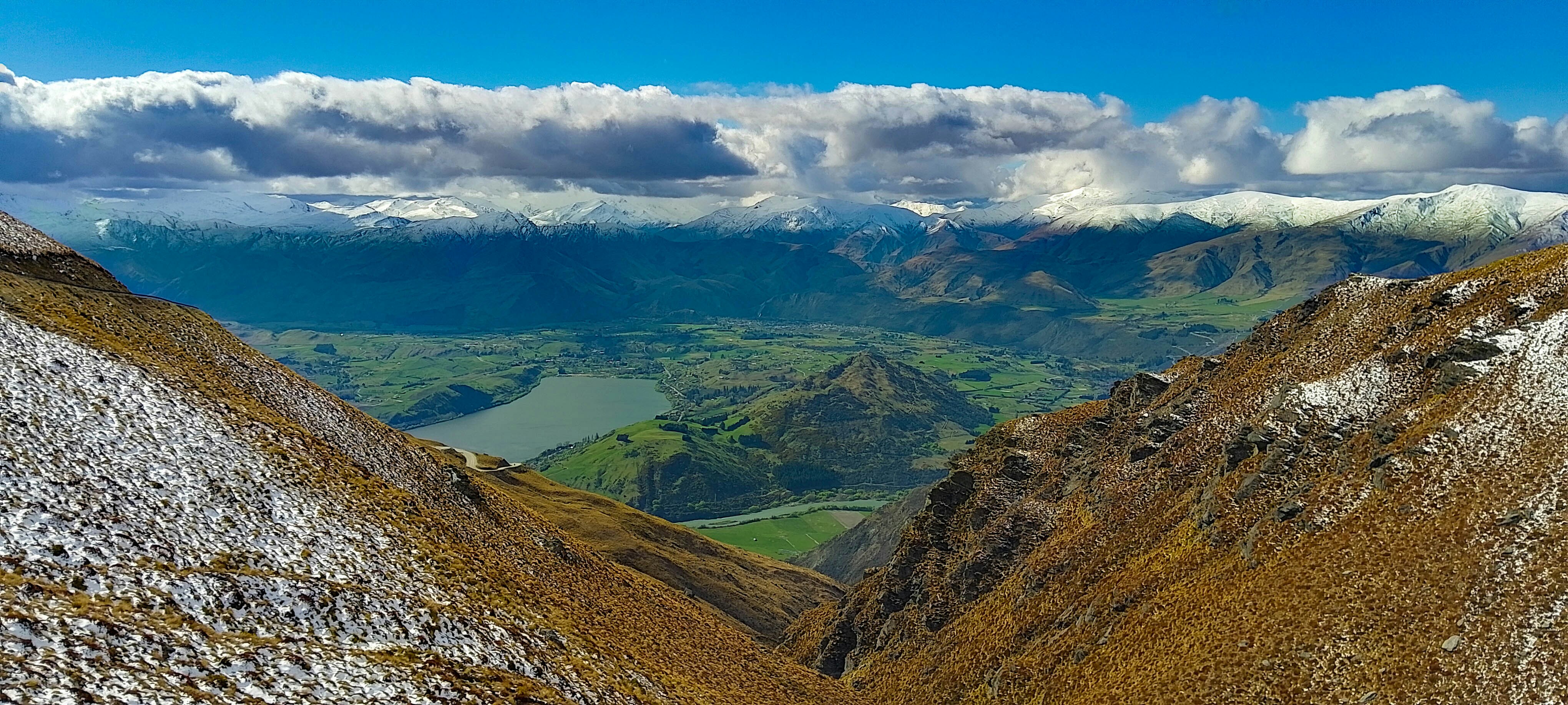 A valley, with mountains surrounding it and big, rolling clouds filling the blue sky. The middle of the valley is quite lush and green, but the mountains are yellowed and snow-covered.