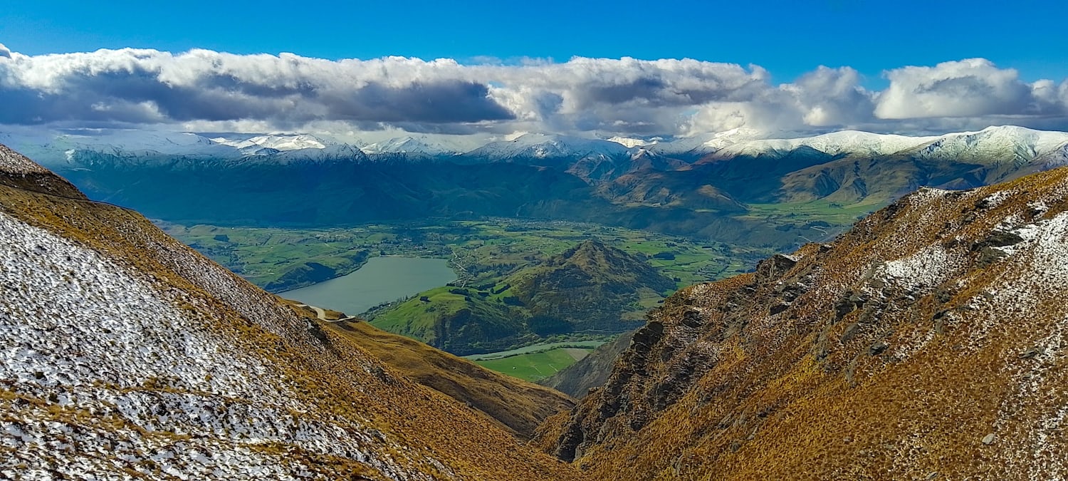 A valley, with mountains surrounding it and big, rolling clouds filling the blue sky. The middle of the valley is quite lush and green, but the mountains are yellowed and snow-covered.