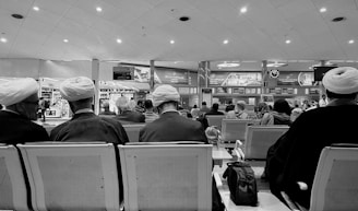 A busy airport waiting area filled with many people sitting on rows of seats. Several individuals are wearing traditional head coverings. Overhead signage includes various advertisements and flight information displays. The scene captures a moment of stillness amidst the bustling environment.