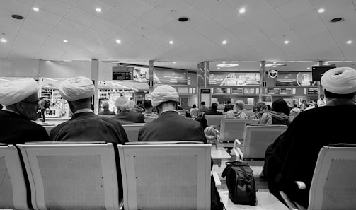 A busy airport waiting area filled with many people sitting on rows of seats. Several individuals are wearing traditional head coverings. Overhead signage includes various advertisements and flight information displays. The scene captures a moment of stillness amidst the bustling environment.