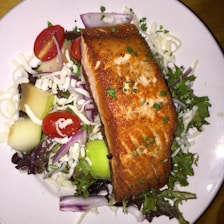 A plate with a grilled salmon fillet placed on a bed of mixed greens, shredded cheese, cherry tomatoes, cucumber slices, and red onion. The salmon is garnished with herbs and appears golden brown and crispy skin.