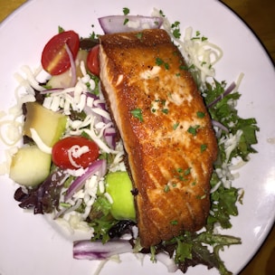 A plate with a grilled salmon fillet placed on a bed of mixed greens, shredded cheese, cherry tomatoes, cucumber slices, and red onion. The salmon is garnished with herbs and appears golden brown and crispy skin.