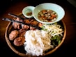 A traditional Vietnamese meal served on a woven bamboo tray, including grilled meat skewers, round meat patties, a heap of white rice noodles, and fresh vegetables like bean sprouts and lettuce. A light-colored bowl contains a sauce with diced carrots and onions. Another small bowl holds sliced green and red peppers.