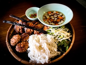 A traditional Vietnamese meal served on a woven bamboo tray, including grilled meat skewers, round meat patties, a heap of white rice noodles, and fresh vegetables like bean sprouts and lettuce. A light-colored bowl contains a sauce with diced carrots and onions. Another small bowl holds sliced green and red peppers.