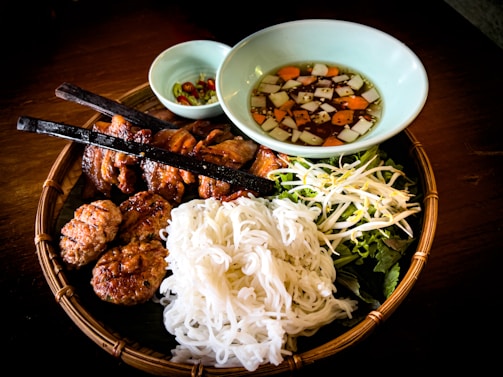 A traditional Vietnamese meal served on a woven bamboo tray, including grilled meat skewers, round meat patties, a heap of white rice noodles, and fresh vegetables like bean sprouts and lettuce. A light-colored bowl contains a sauce with diced carrots and onions. Another small bowl holds sliced green and red peppers.