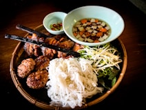A traditional Vietnamese meal served on a woven bamboo tray, including grilled meat skewers, round meat patties, a heap of white rice noodles, and fresh vegetables like bean sprouts and lettuce. A light-colored bowl contains a sauce with diced carrots and onions. Another small bowl holds sliced green and red peppers.