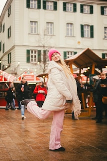 A person in a white winter jacket and pink pants, with a vibrant pink beanie, poses playfully with one leg raised in a festive outdoor market setting. The background features a historic building with green shutters and various market stalls adorned with lights. Other people, dressed warmly, are visible, creating a lively and cheerful atmosphere.