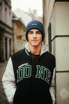 A young person is leaning against an urban wall, wearing a blue beanie labeled with 'GUESS' and a jacket with the word 'DONE'. The background features architectural details of buildings and a narrow street.