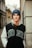 A young person is leaning against an urban wall, wearing a blue beanie labeled with 'GUESS' and a jacket with the word 'DONE'. The background features architectural details of buildings and a narrow street.