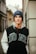A young person is leaning against an urban wall, wearing a blue beanie labeled with 'GUESS' and a jacket with the word 'DONE'. The background features architectural details of buildings and a narrow street.