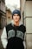 A young person is leaning against an urban wall, wearing a blue beanie labeled with 'GUESS' and a jacket with the word 'DONE'. The background features architectural details of buildings and a narrow street.