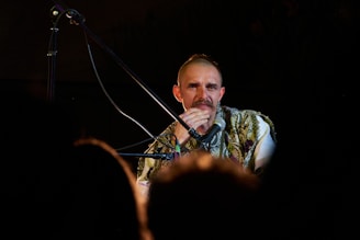 A person is seated in a dimly lit environment, holding a microphone. The individual appears to be contemplating or engaged in deep thought, with a focused gaze. The person is wearing a patterned vest over a white shirt. There are shadows and silhouettes of people or objects in the foreground, suggesting a live performance or public speaking event.