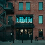 A brick building features a café with a sign that reads 'The Coffee House' above a black awning. The building has large windows with a teal tint and an external staircase on the left side. Two chairs and a table are set outside.