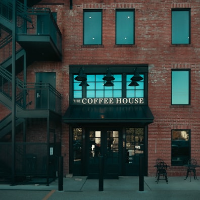 A brick building features a café with a sign that reads 'The Coffee House' above a black awning. The building has large windows with a teal tint and an external staircase on the left side. Two chairs and a table are set outside.