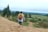 A person with a backpack walks barefoot on a sandy path, holding their shoes. The landscape includes green fields and trees under an overcast sky.