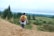 A person with a backpack walks barefoot on a sandy path, holding their shoes. The landscape includes green fields and trees under an overcast sky.