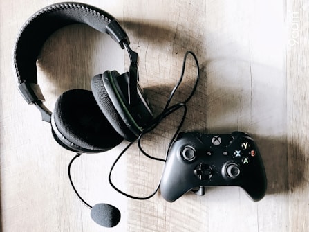 A black gaming headset with a microphone is placed next to a black game controller on a wooden surface. The headset features padded ear cups and a headband, while the controller has colored buttons for easy identification.