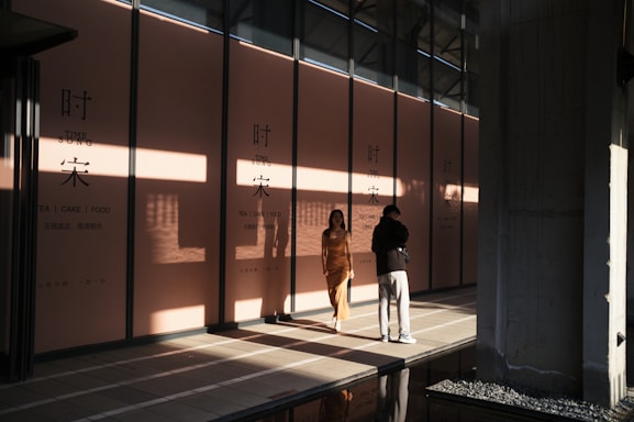 Two people stand in front of a large storefront with glass walls and high ceiling. The sunlight creates shadows and highlights on the peach-colored panels with signage that reads 'TIME SONG' and 'TEA | CAKE | FOOD'. The atmosphere is modern and serene, with reflections on the tiled floor.