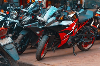 A collection of parked motorcycles, featuring a prominent red and black sport bike in the foreground. The image shows multiple bikes closely lined up, each with varying designs and colors, primarily dominated by shades of black and red. The setting appears to be urban, possibly a market area or street.