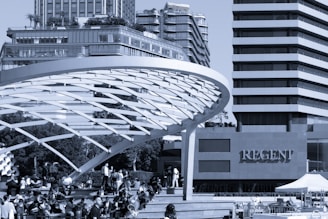 A modern urban scene with a large, curved architectural structure in the foreground. The structure provides shade for numerous people gathered on steps below, some sitting and others walking. In the background, high-rise buildings tower above, featuring glass facades and tiered balconies. The word 'Regent' is prominently displayed on one of the buildings.