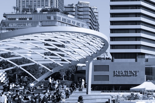 A modern urban scene with a large, curved architectural structure in the foreground. The structure provides shade for numerous people gathered on steps below, some sitting and others walking. In the background, high-rise buildings tower above, featuring glass facades and tiered balconies. The word 'Regent' is prominently displayed on one of the buildings.