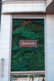 A vertical garden is integrated into the facade of a building, featuring lush green foliage. A rectangular sign with white script is centrally positioned on the garden wall. The exterior is made of light-colored stone tiles, complementing the natural green hues of the plants.