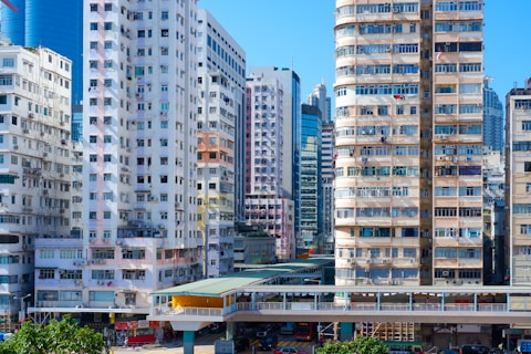 A densely packed urban landscape features tall residential and commercial buildings with various architectural styles. The structures are tightly arranged, and many have balconies with items like air conditioning units and laundry. A pedestrian bridge or walkway connects parts of the cityscape. The overall scene is vibrant, bustling, and indicative of a busy metropolitan area under clear blue skies.