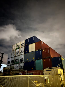A row of various sizes of shipping containers stacked neatly in a dimly lit industrial yard.