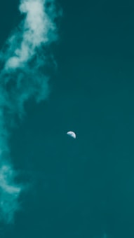 The image features a vibrant, teal sky with scattered white clouds, highlighted by a visible half-moon near the center. The contrast between the moon and the sky provides a tranquil and serene atmosphere.