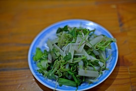 A blue and white plate is filled with fresh, leafy greens and stalks. The dish appears to be raw, showcasing a variety of green vegetables that are loosely arranged on the plate.