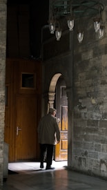 An elderly person, dressed in neutral clothing, is seen standing next to an open wooden door in a dimly lit, rustic interior with stone walls. The scene is illuminated by natural light streaming through the doorway, creating a stark contrast between the dark and light areas. Hanging lanterns are visible above, and wooden panels line one side of the wall.