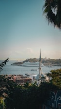 A scenic view of a coastal city with a prominent mosque in the foreground. The skyline includes a large cruise ship on the water, and a mix of historic and modern buildings. Lush greenery and palm fronds frame the scene.