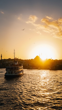 A scenic view of a ferryboat on a body of water during sunset. The sun is setting on the horizon, filling the sky with a golden hue. Silhouettes of buildings and minarets are visible in the background, while a few birds are flying in the sky.