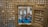Viewed through a latticed frame, the interior of a grand hall features a speaker at a podium, clad in traditional attire. The blue carpet contrasts with the stone columns and high windows, while an audience sits attentively.