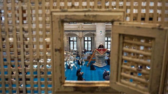Viewed through a latticed frame, the interior of a grand hall features a speaker at a podium, clad in traditional attire. The blue carpet contrasts with the stone columns and high windows, while an audience sits attentively.
